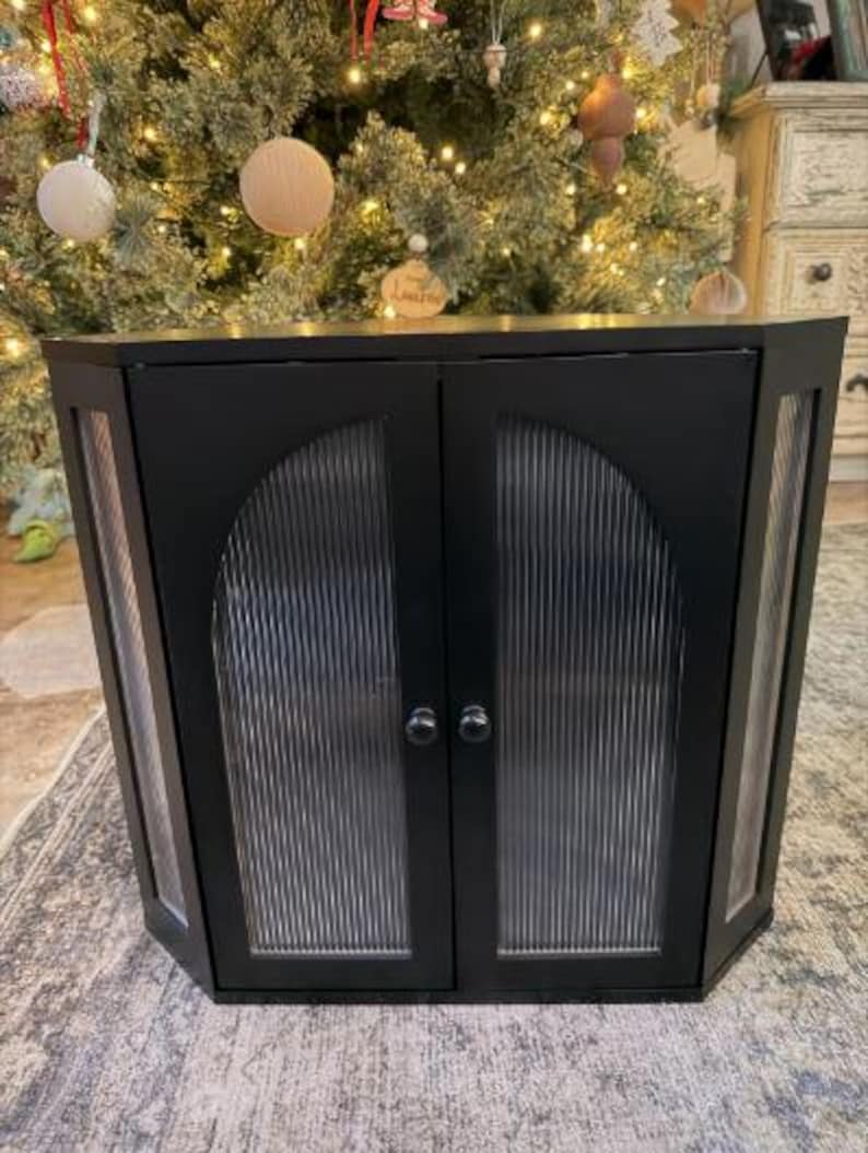 May include: A black corner cabinet with two doors and glass panels. The cabinet has a modern design with a curved top and silver hardware. The glass panels have a vertical ribbed texture. The cabinet is placed in front of a Christmas tree.