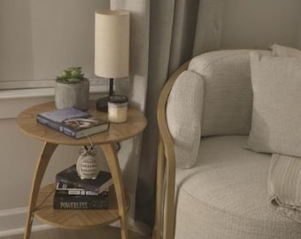 Könnte beinhalten: Eine gemütliche Leseecke mit einem runden Beistelltisch aus Holz, Büchern, einer kleinen Lampe und einer Sukkulente. Ein cremefarbener Sessel und neutrale Vorhänge schaffen eine entspannte Atmosphäre. Der Tisch hat etwa 60 cm Durchmesser.
