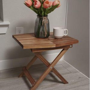 May include: A small, folding wooden side table with a natural finish. It holds a glass vase with pink tulips and a white mug. The table is set against a light-coloured wall and floor, creating a simple, inviting scene.