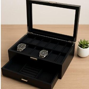 May include: A black wooden watch box with a clear glass top, open to reveal slots for watches. Two watches are displayed. The bottom drawer is open, revealing compartments for additional storage. A small potted plant sits to the side.