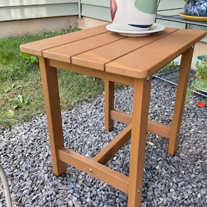 Puede incluir: Una mesa auxiliar rectangular marrón con una parte superior de listones. La mesa está hecha de material similar a la madera y tiene cuatro patas. Un plato blanco con una maceta decorativa se encuentra encima. La mesa mide aproximadamente 61 cm de alto.