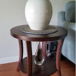 May include: A dark brown, round side table with a lower shelf and curved legs. A white ceramic lamp with a white lampshade sits on top. A decorative glass vase is on the lower shelf. The table is 61 cm in diameter.