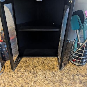 May include: Black corner cabinet with two doors ajar, revealing a shelf inside. The doors have a metal mesh design. The cabinet is on a countertop with a speckled pattern. A yellow container sits on top.