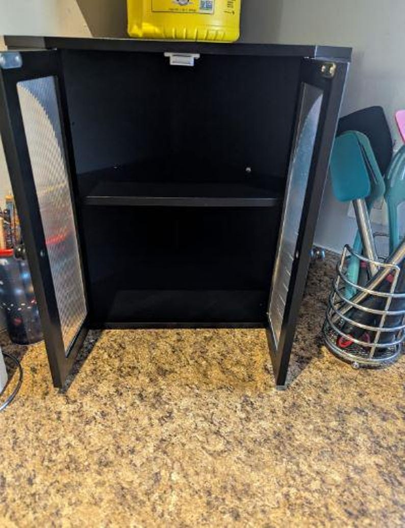 May include: Black corner cabinet with two doors ajar, revealing a shelf inside. The doors have a metal mesh design. The cabinet is on a countertop with a speckled pattern. A yellow container sits on top.