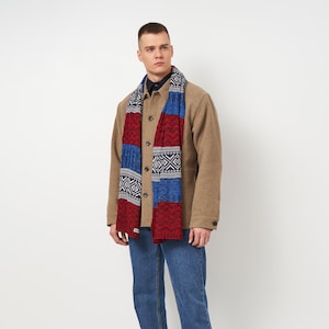 May include: A patterned scarf in red, blue, and white is draped around the neck. The scarf features geometric designs and is worn over a tan jacket and blue jeans. The scarf is a winter accessory.