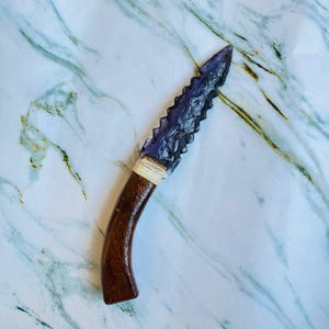 May include: A handcrafted knife with a dark purple, jagged blade and a curved, wooden handle. The blade is attached to the handle with a wrapped, beige material. The knife is displayed on a white and grey marble surface.