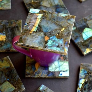 May include: Square labradorite coasters with a purple teacup. The coasters are made of a mosaic of blue, green, and gold-colored stones. The teacup is resting on one of the coasters. Other coasters are scattered around the teacup.