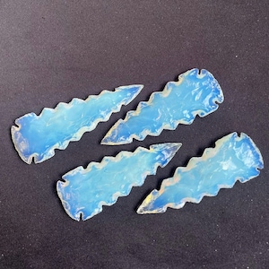 May include: Four opalescent, blue-white gemstone arrowheads with jagged edges. The translucent stones have a subtle, ethereal glow, and are arranged on a dark background. The arrowheads are a unique, handcrafted item.
