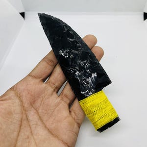 May include: A black obsidian knife with a yellow wrapped handle. The blade is a dark, glossy black, and the handle is wrapped with bright yellow thread. The knife is held in a hand.