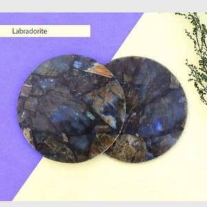 May include: Two round Labradorite coasters with a mix of blue, brown, and gray tones. The coasters are displayed on a background of purple and cream colors, with a sprig of greenery in the corner. The word "Labradorite" is in a white rectangle.