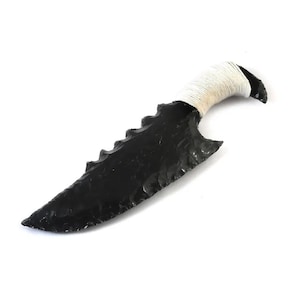 May include: A black obsidian knife with a serrated edge and a wrapped white handle. The blade has a pointed tip and a curved shape. The handle is tightly wrapped with a white cord. The knife is set against a white background.