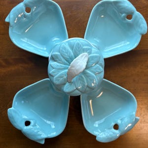May include: A light blue ceramic serving dish set. The set includes four individual dishes, each with a unique shape and a small hole, surrounding a central, decorative piece. The central piece has a leaf design and a white accent.