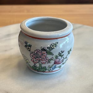 Puede incluir: Pequeño jarrón redondo de cerámica blanca con diseño floral. El jarrón presenta flores rosas y blancas con hojas verdes y un borde rojo. El jarrón está sobre una superficie de mármol blanco.