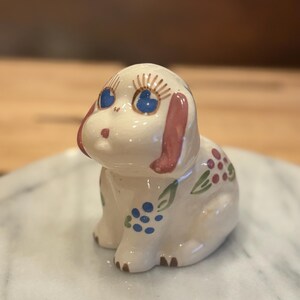 May include: A ceramic dog figurine with a cream-colored body, blue eyes, and pink ears. The figurine is decorated with blue and red floral patterns and brown paws. The dog is sitting and has a sweet, innocent expression.
