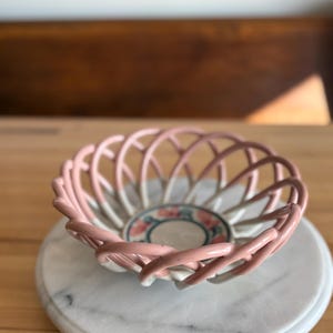 May include: A pink ceramic basket with a lattice design, resting on a white marble coaster. The basket's interior features a floral pattern in pink and green. The basket is a decorative item.