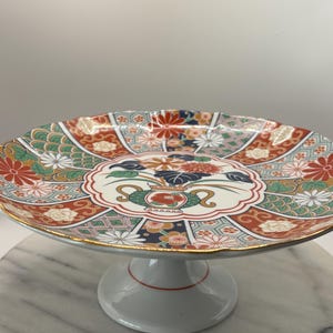 Puede incluir: Un plato de pedestal de cerámica decorativo con un diseño floral colorido. El plato presenta una base blanca con una línea de acento roja, un borde dorado y un patrón detallado de flores en rojo, verde y blanco. El plato está elevado sobre un pedestal blanco.