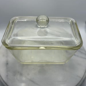 Könnte beinhalten: Eine klare, quadratische Glasschale mit passendem Deckel. Der Deckel hat einen kleinen, runden Knopf zum einfachen Anfassen. Die Schale steht auf einer weißen und grauen Marmoroberfläche. Das Glas hat einen leichten Gelbstich.