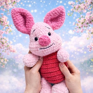 Peut inclure: Une peluche au crochet faite à la main représentant un personnage rose avec de grandes oreilles, un nez rond et une chemise rayée rouge. Le jouet est présenté sur un fond de ciel bleu avec des nuages blancs et des fleurs roses.