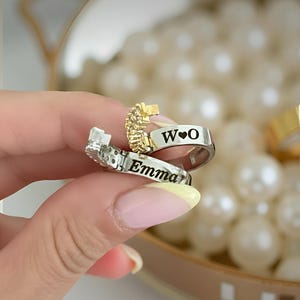 May include: A personalized silver ring with the name "Emma" engraved on it, and a gold ring with the initials "W❤O". The rings are set against a backdrop of white pearls.