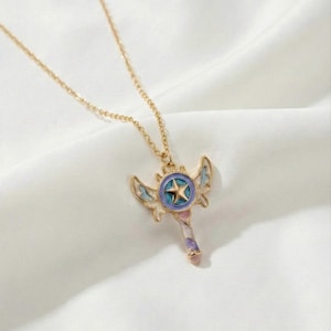 May include: A gold-coloured necklace with a pendant shaped like a star-topped wand with wings. The pendant has a blue, purple, and pink colour scheme. The necklace is displayed on a white, textured surface.