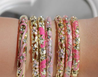 Könnte beinhalten: Ein Set aus sieben Armreifen aus goldfarbenem Metall mit floralen Mustern aus rosa und weißem Emaille sowie eingelegten klaren Kristallen. Die Armreifen zeigen filigrane Ausschnittdesigns und verschiedene Rosatöne im Emaille.