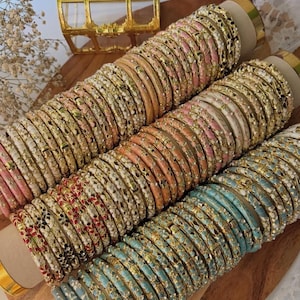 Puede incluir: Un surtido de pulseras coloridas en tonos rosa, naranja y azul, con detalles metálicos dorados. Las pulseras están dispuestas en filas ordenadas, mostrando intrincados patrones florales y geométricos. Una pequeña y ornamentada caja de joyería dorada se encuentra en el fondo.