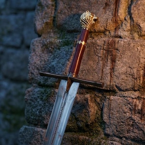 Pode incluir: Uma espada longa com lâmina metálica apresentando um sulco central, um punho castanho-avermelhado escuro e uma guarda reta. O pomo tem a forma de uma cabeça de leão dourada detalhada, com uma fina faixa dourada no topo do cabo.