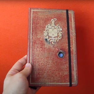 Puede incluir: Un diario de estilo vintage con una cubierta roja desgastada, un emblema dorado y un cierre elástico negro. El diario tiene un detalle azul en forma de botón y se sostiene sobre un fondo naranja.