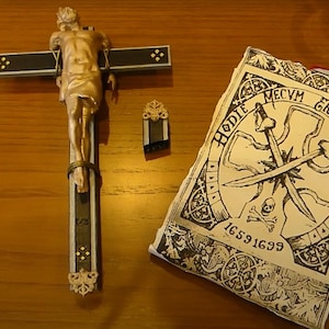 Puede incluir: Un crucifijo dorado con una figura detallada de Jesús, una pequeña pieza decorativa negra y dorada, y un pergamino de estilo vintage con texto y espadas cruzadas. El pergamino dice "Hodie Mecum Eris In Paradiso" y tiene el número "16591699".