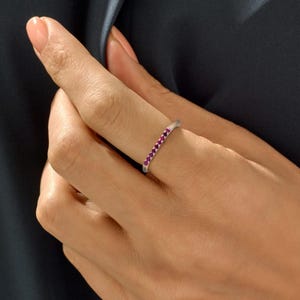 May include: A silver ring set with a line of small, bright pink gemstones. The ring is shown on a finger, highlighting its delicate design and the sparkle of the stones. The ring's band is slender and polished, accentuating the vibrant gemstones.
