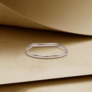 May include: A silver ring with a rectangular top and a rounded band. The ring is set against a neutral background, highlighting its simple, elegant design. The ring's polished surface reflects light, showcasing its metallic sheen.