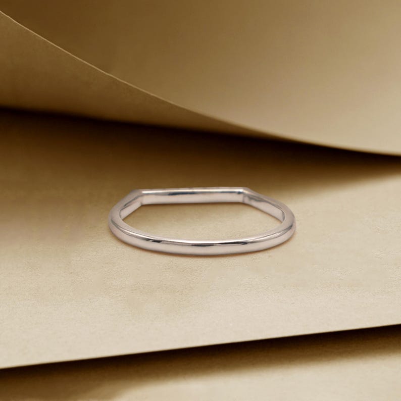 May include: A silver ring with a rectangular top and a rounded band. The ring is set against a neutral background, highlighting its simple, elegant design. The ring's polished surface reflects light, showcasing its metallic sheen.