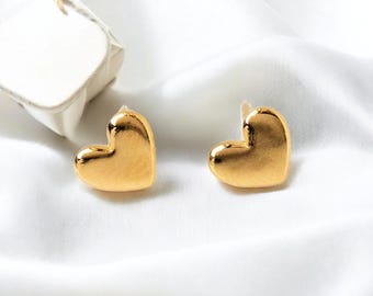 May include: Two gold-colored heart-shaped stud earrings. The earrings are on a white satin surface, with a small white jewelry box in the background. The earrings have a shiny, reflective surface.
