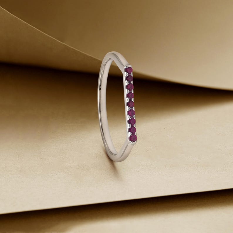 May include: A silver ring featuring a line of small, deep red gemstones. The ring has a minimalist design with a slender band and a polished surface. The gemstones are evenly spaced, providing a vibrant contrast. The ring is a piece of jewellery.