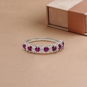 May include: A silver ring featuring alternating round, deep red gemstones and smaller, clear gemstones. The ring is set against a neutral background, with a partially visible jewelry box in the background.