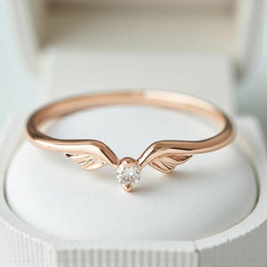 May include: Rose gold ring with a delicate wing design and a small, clear gemstone at the center. The ring is set against a white background, showcasing its elegant details. The wings curve upwards, adding a unique touch to the jewelry.