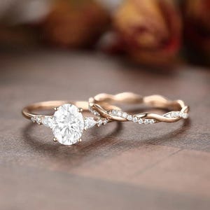 Puede incluir: Un juego de anillos de compromiso de oro rosa. El anillo de compromiso presenta una piedra central de talla ovalada flanqueada por piedras laterales en forma de pera. La banda a juego tiene un diseño retorcido con pequeñas piedras brillantes.