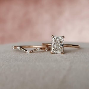 May include: Two gold rings. One ring features a large, rectangular, clear gemstone. The other ring is a curved band with small, clear gemstones. The rings are set against a soft, pink background.