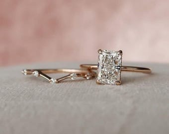 May include: Two gold rings. One ring features a large, rectangular, clear gemstone. The other ring is a curved band with small, clear gemstones. The rings are set against a soft, pink background.