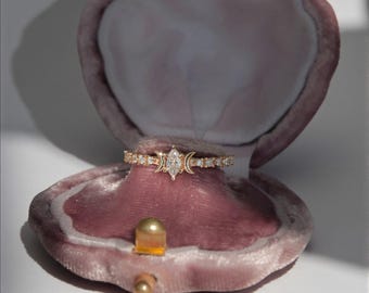 May include: A gold ring with a marquise-cut diamond center stone, flanked by crescent moon and small diamond accents. The ring is displayed in a pink velvet jewelry box with a gold clasp.