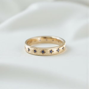 Peut inclure: Une bague en or ornée de motifs en forme d'étoiles, chacun contenant une petite pierre précieuse bleu foncé. La bague est présentée sur un fond blanc doux, soulignant sa surface polie et ses détails complexes. Le design de la bague est symétrique et élégant.