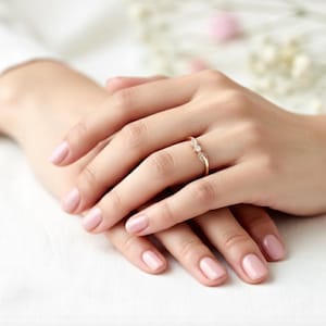May include: A delicate gold ring with a small, clear gemstone is worn on a finger. The ring is set against a background of hands with manicured nails. The nails are painted a soft pink color. The ring is the focal point of the image.