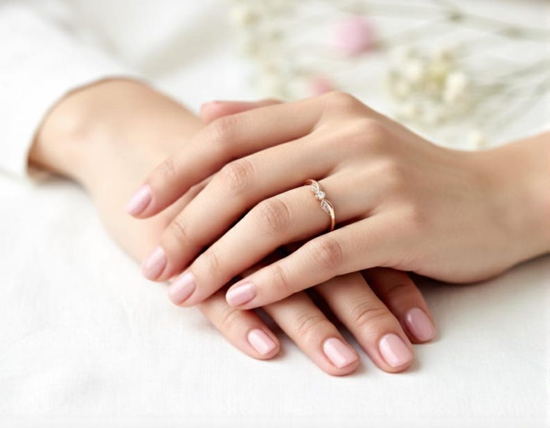 May include: A delicate gold ring with a small, clear gemstone is worn on a finger. The ring is set against a background of hands with manicured nails. The nails are painted a soft pink color. The ring is the focal point of the image.