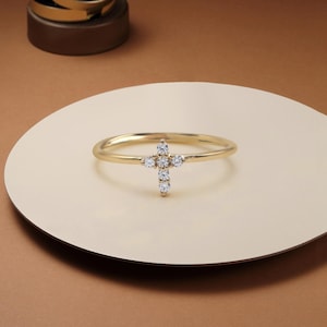 Peut inclure: Une bague dorée ornée d'une croix sertie de petites pierres précieuses transparentes. La bague est posée sur une surface crème, avec un fond marron. Un bijou délicat et élégant.