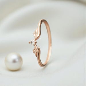 May include: Rose gold ring featuring a leaf design with a small, clear gemstone at the center. The ring has a delicate, curved band and two stylized leaf accents. A pearl is in the background.