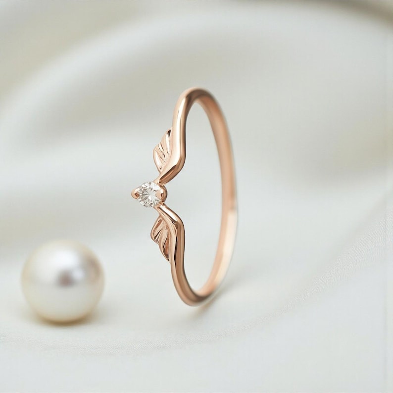 May include: Rose gold ring featuring a leaf design with a small, clear gemstone at the center. The ring has a delicate, curved band and two stylized leaf accents. A pearl is in the background.