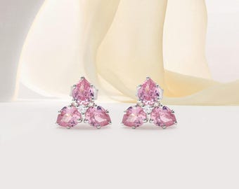 May include: A pair of silver stud earrings, each featuring three pink pear-shaped gemstones and a small white gemstone. The earrings are set against a soft, neutral background.