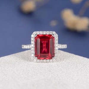 Puede incluir: Un anillo de plata con una gran gema rectangular de color rojo intenso, rodeada por un halo de gemas más pequeñas y transparentes. La banda del anillo también está adornada con pequeñas gemas transparentes. El anillo se encuentra sobre una superficie blanca.
