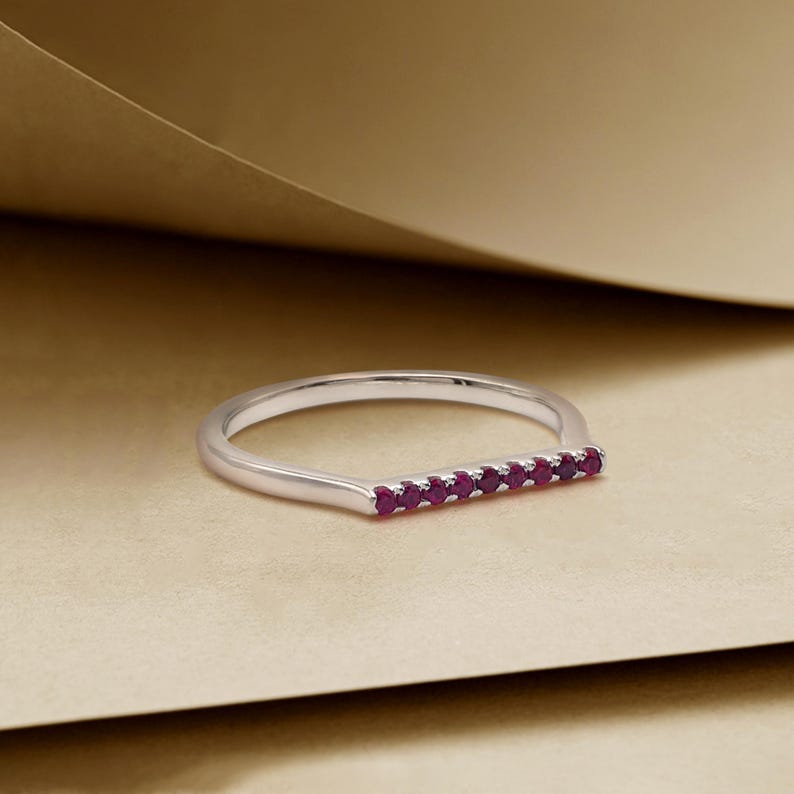 May include: A silver ring featuring a line of small, round, red gemstones. The ring is displayed on a neutral surface, emphasizing the design and colour of the jewellery.