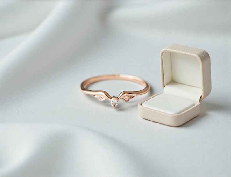 May include: A rose gold ring with a wing design and a central clear gemstone, next to an open beige ring box. The ring is set against a white, textured background, creating a soft, elegant aesthetic.